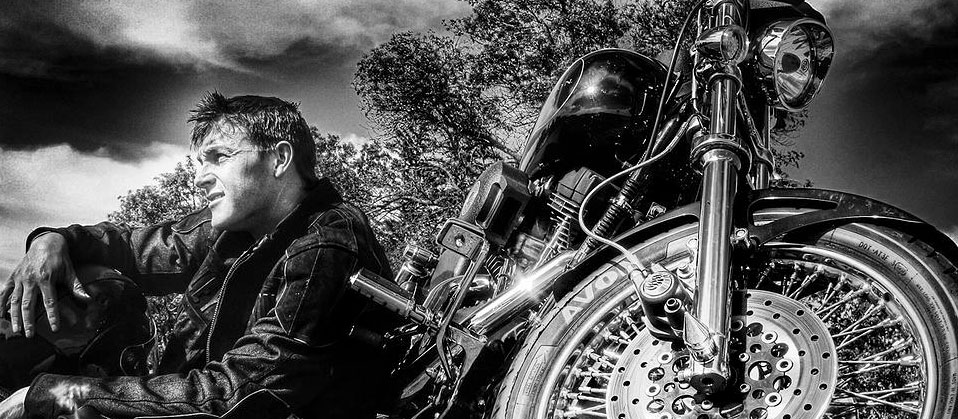 black and white art photo of a man seated on the ground next to a motorcycle