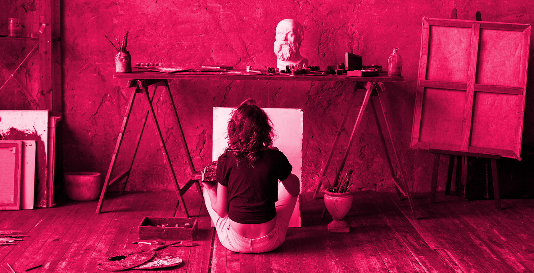 red stained photo of young woman seated on the floor of studio painting view from behind